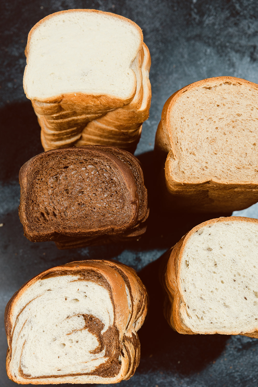 Stacked cross-section slices of La Segunda basket breads on a dark surface — white, wheat, marble, and pumpernickel — showing the classic square pan-loaf shape and crumb of each variety.