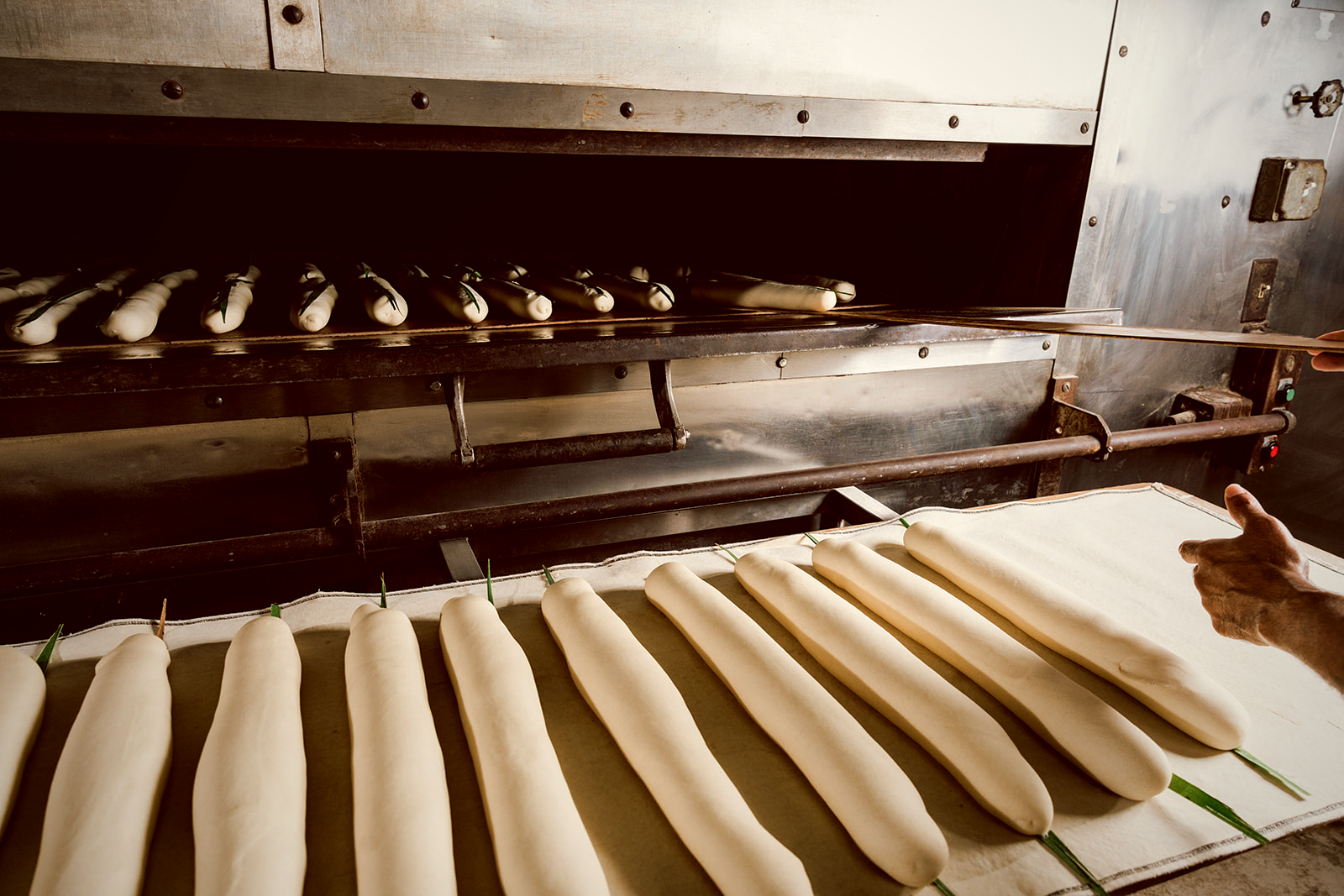 A baker loads shaped Cuban loaves into the long stone-hearth oven with a wooden peel, leafed loaves waiting on the canvas board in the foreground.