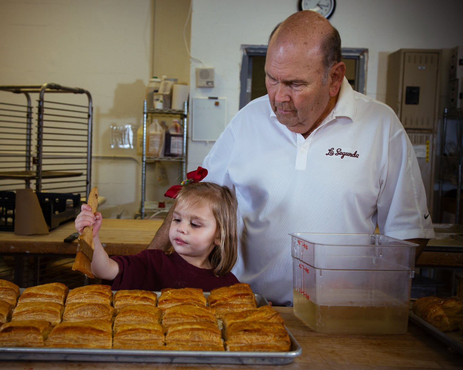 Tony Moré in a La Segunda polo watching his granddaughter glaze a tray of fresh pastries on the bakery floor.