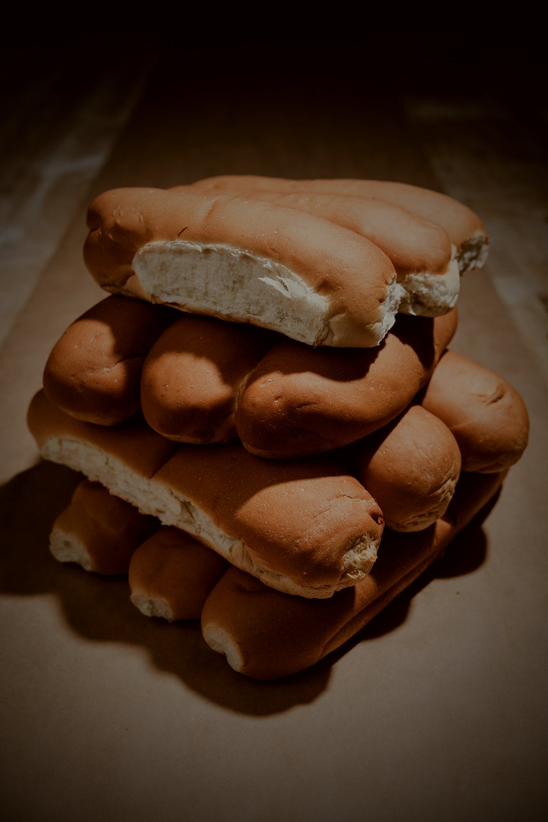 A stack of fresh 8-inch and 12-inch hoagie rolls in soft warm bakery light against a deep shadow background.