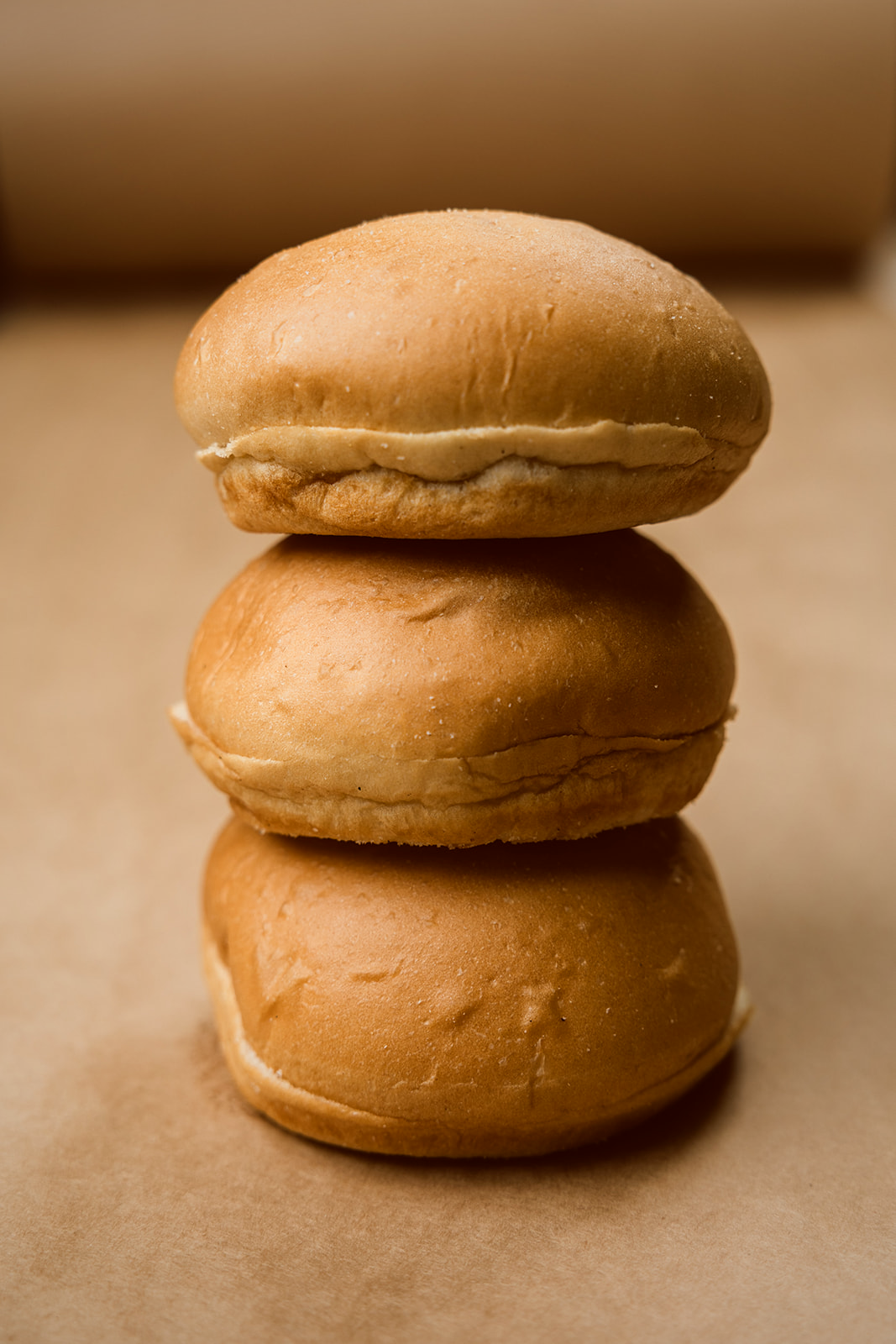 A stack of three Coca hamburger buns shown from the side on warm parchment, the pre-sliced cut visible between them and a glossy pillowy crown on top.
