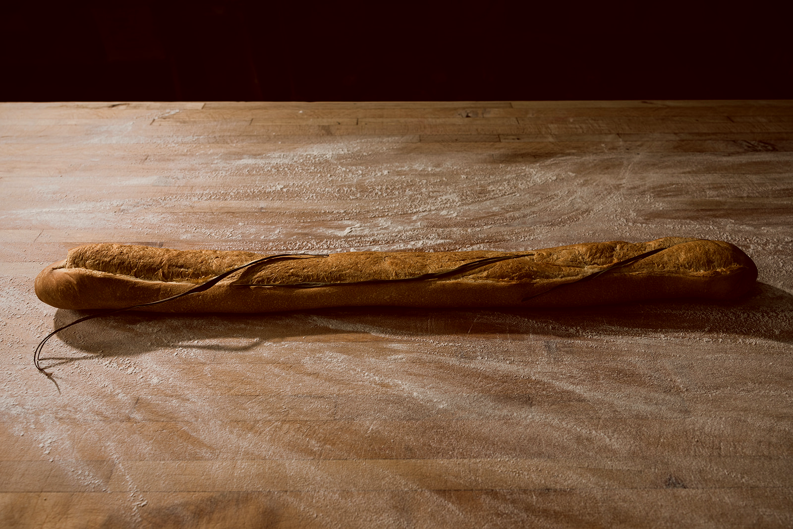 A full 36-inch La Segunda Cuban bread loaf laid horizontally on a flour-dusted wooden board, the freshly cut palmetto leaf still pressed into the split and trailing off the end.