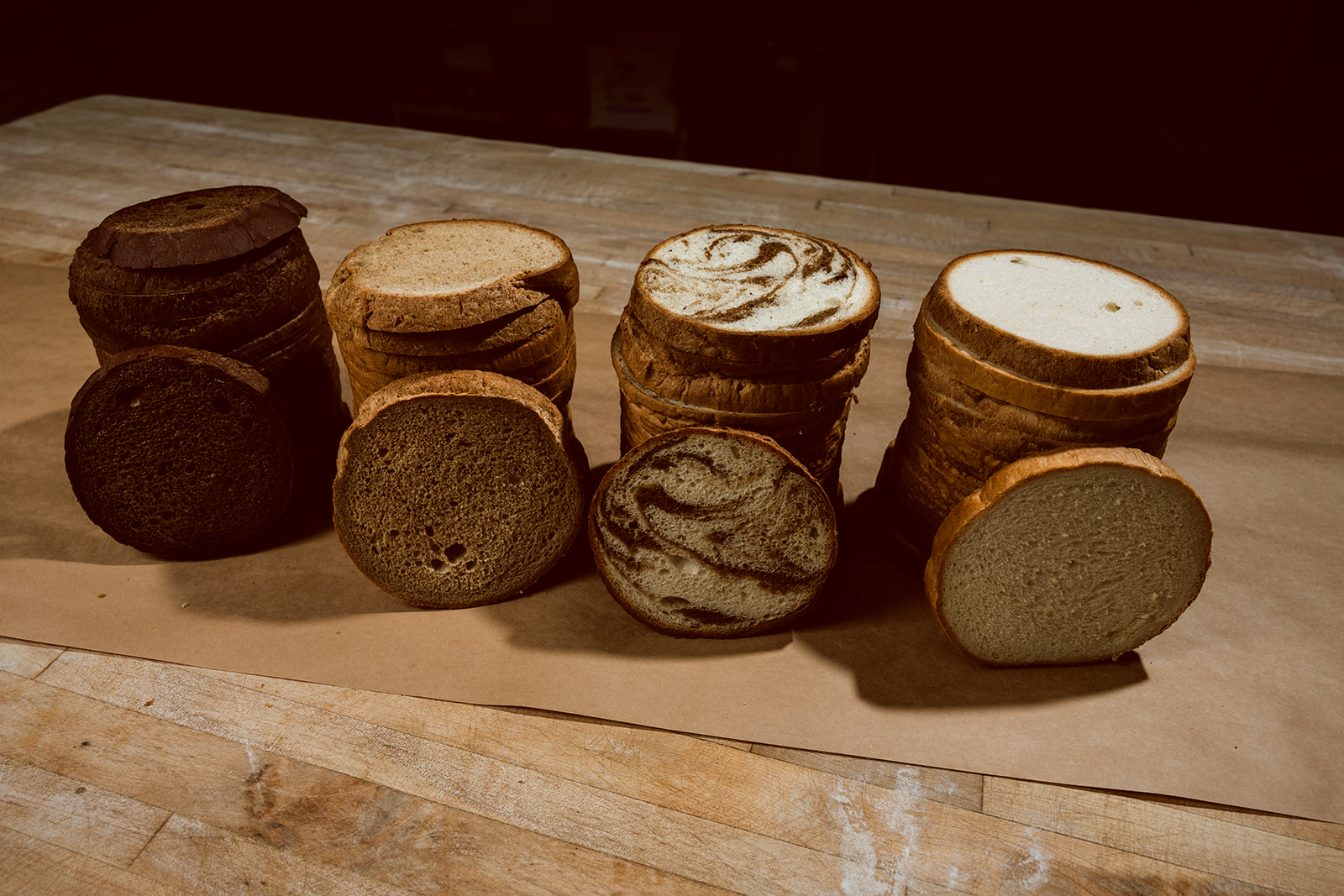 Stacked cross-section slices of La Segunda bucket breads on kraft paper — pumpernickel, rye, marble, and white — showing the distinct crumb and color of each variety.