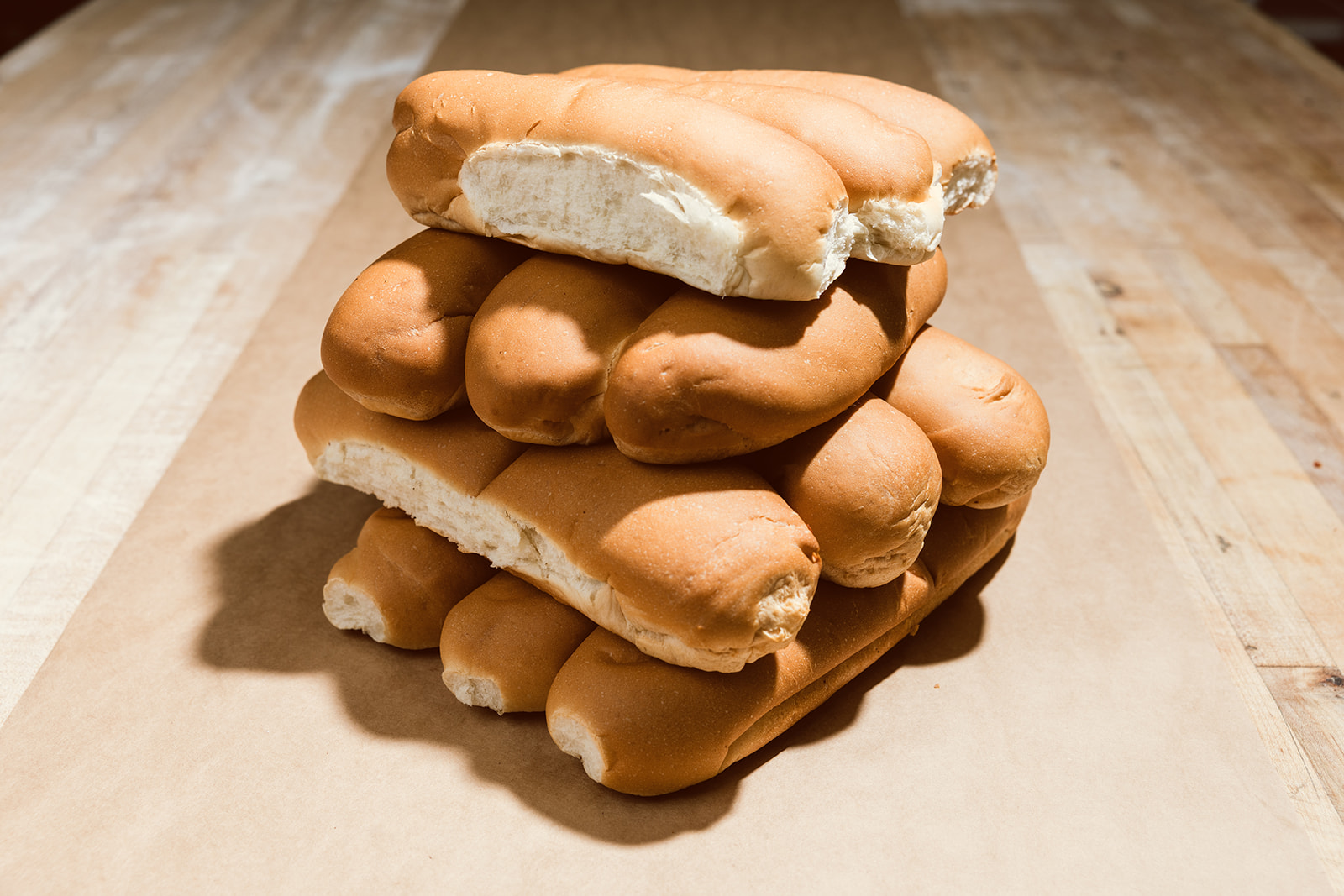 A stacked pyramid of fresh 8-inch La Segunda hoagie rolls on kraft paper, the top roll torn open to reveal its soft white crumb.