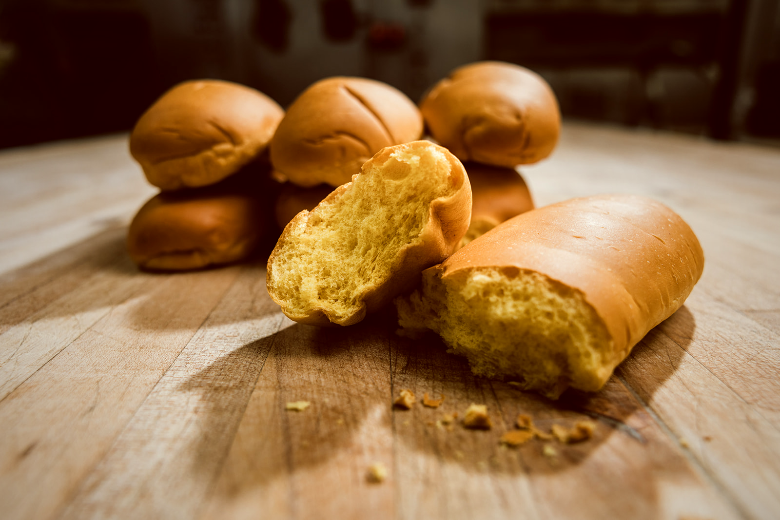 A cluster of golden egg-washed Media Noche hoagie rolls on a warm wooden board, one torn open in the foreground revealing the soft golden crumb, crumbs scattered.